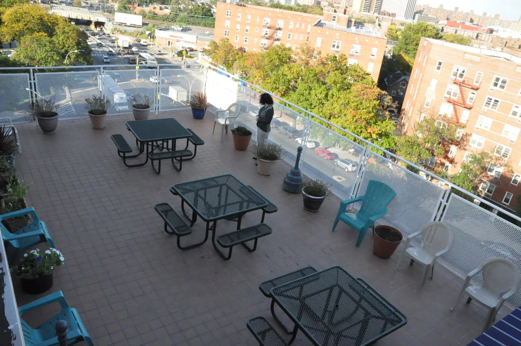 A rooftop patio with several tables and chairs surrounded by potted plants in an urban environment.