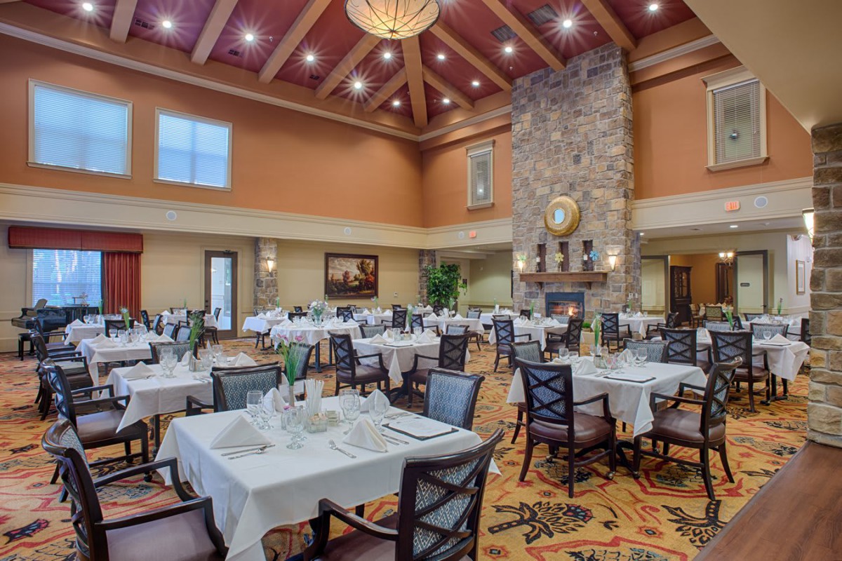 A spacious and elegantly designed dining room with neatly arranged tables covered in white tablecloths, featuring a prominent stone fireplace under decorative lighting.
