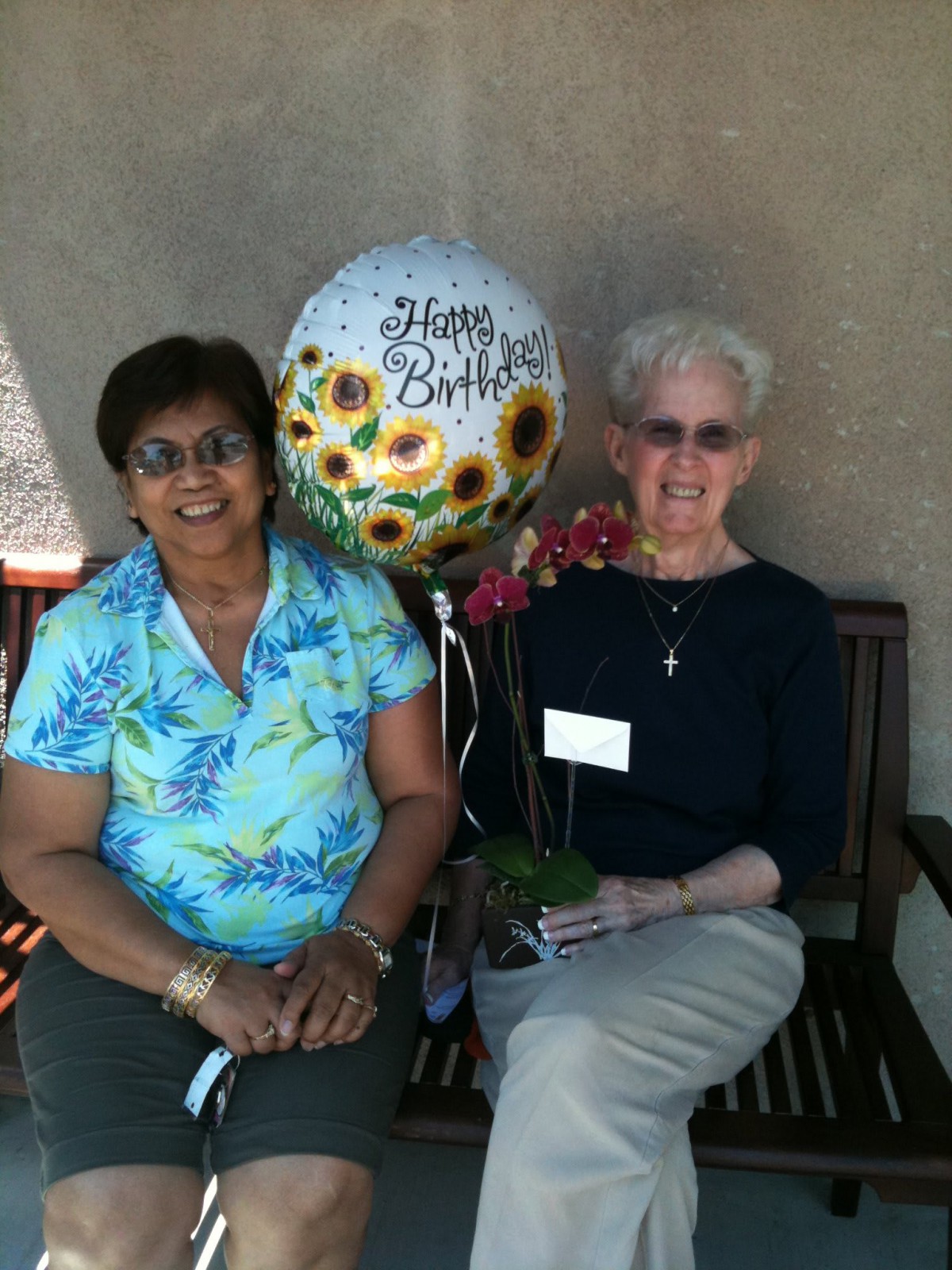 Two women sitting on a bench outdoors, celebrating a birthday with a balloon and flowers.