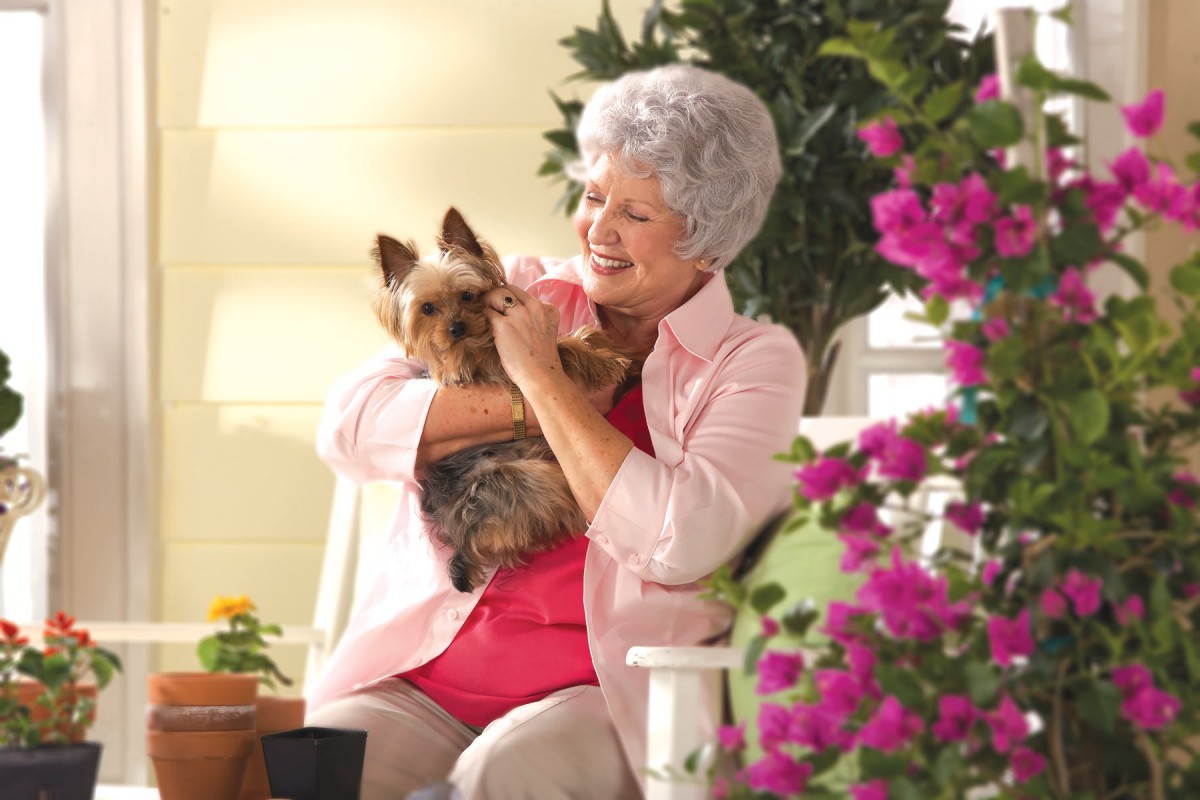 An elderly woman smiling and holding a small dog while surrounded by colorful flowers in a garden.