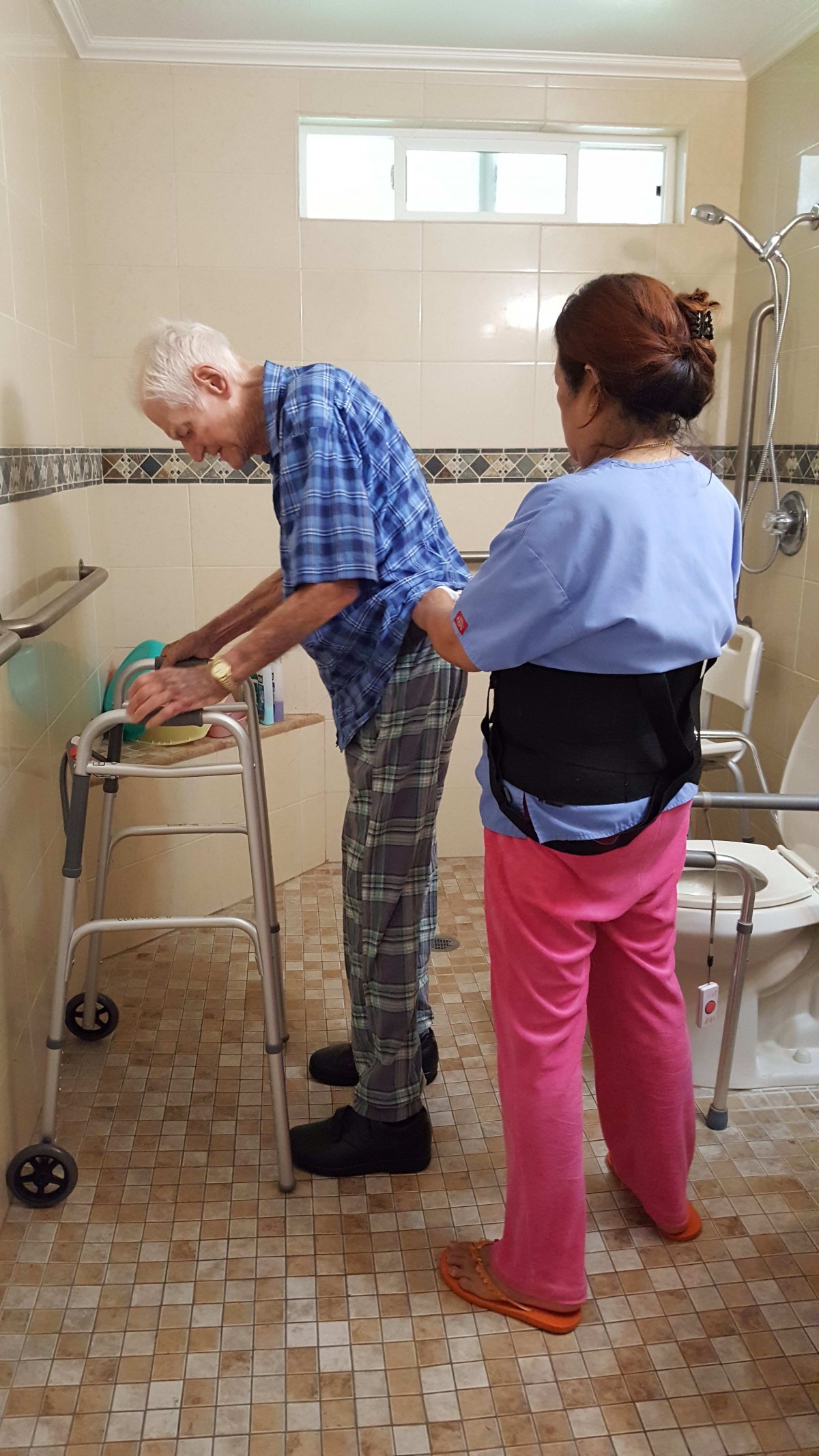 A caregiver assists an elderly man with a walker in a bathroom setting.