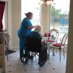 A caregiver assisting an elderly person in a wheelchair at a dining area.
