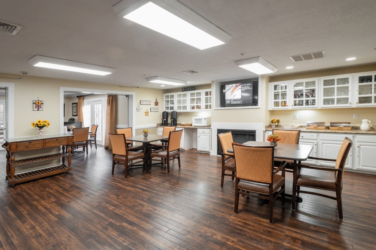 A well-lit dining area with wooden flooring, featuring tables, chairs, cabinets, and a television.