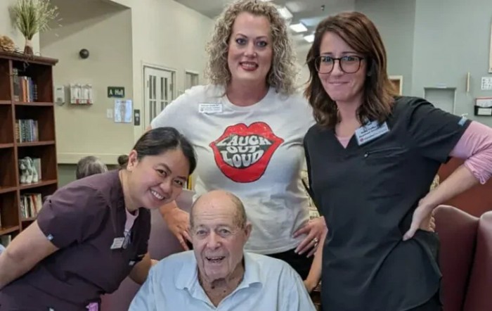 A cheerful group photo featuring an elderly man seated with three women standing behind him, all smiling in a casual indoor setting.