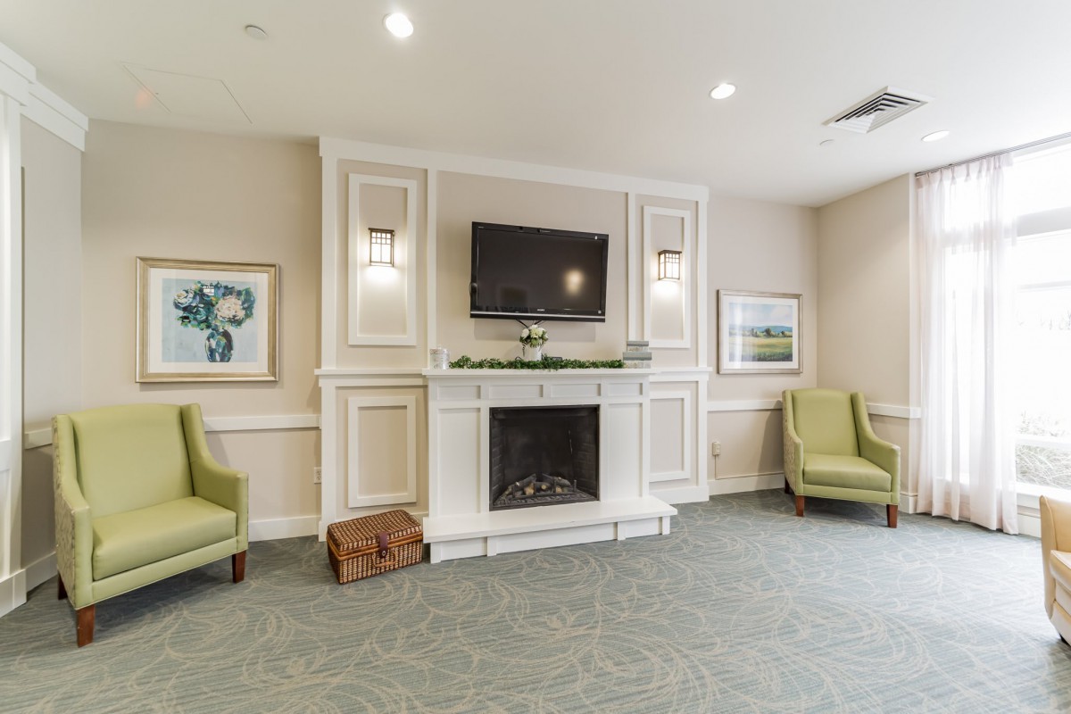 A cozy living room features a fireplace beneath a mounted television, flanked by two comfortable green chairs and tasteful artwork on the walls.