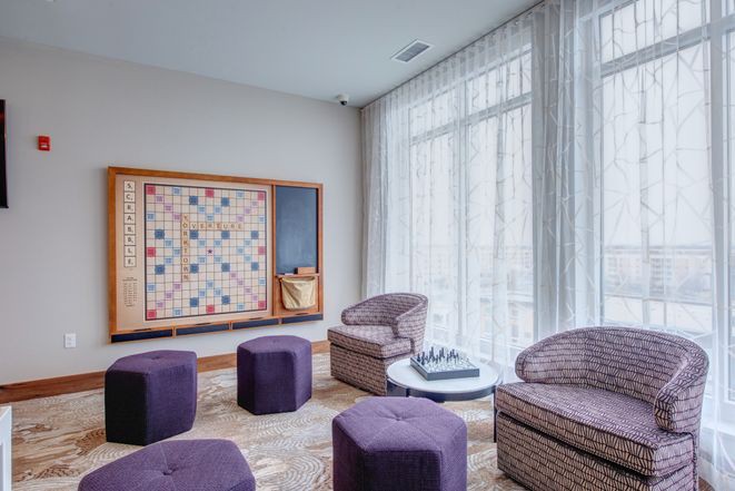 A modern lounge area with purple seating and a chess set, illuminated by natural light from a large window with sheer curtains.