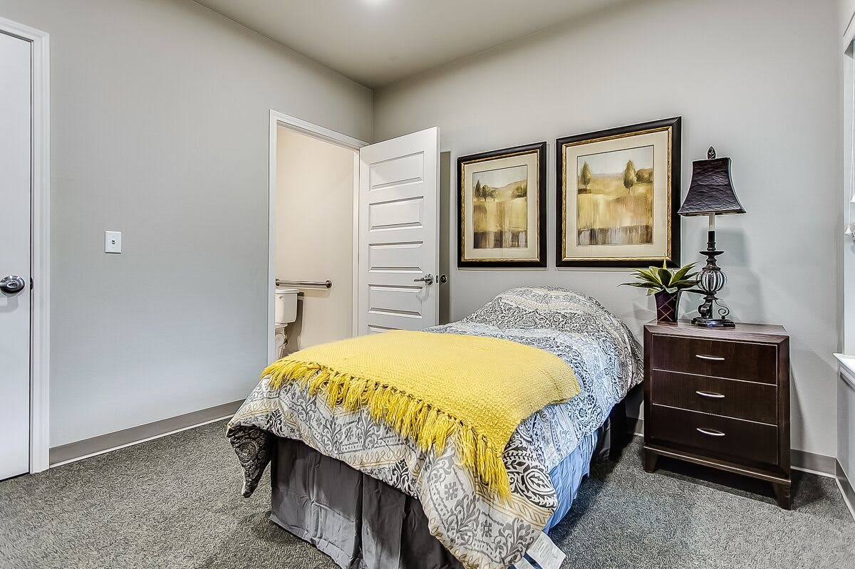 A cozy bedroom featuring a single bed with a yellow blanket, paintings on the wall, and a nightstand with a lamp.