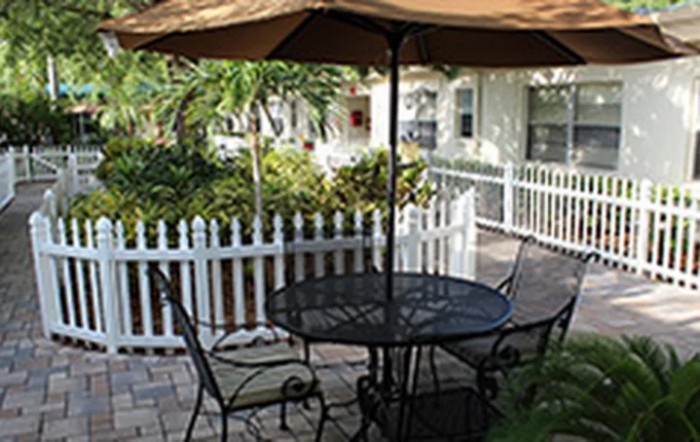 A cozy patio with a metal table and chairs under a large umbrella, surrounded by a white fence and lush tropical plants.