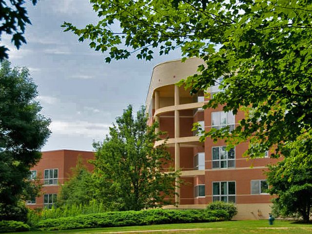 A modern brick building surrounded by lush green trees and shrubs under a cloudy sky.
