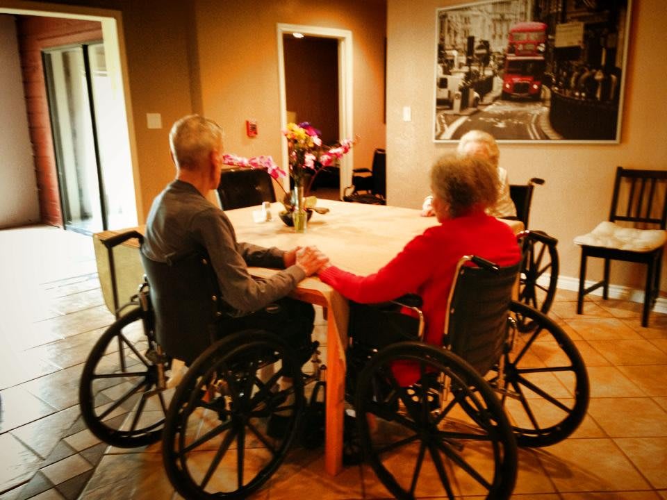 Elderly individuals sitting at a table in wheelchairs within a care home setting.