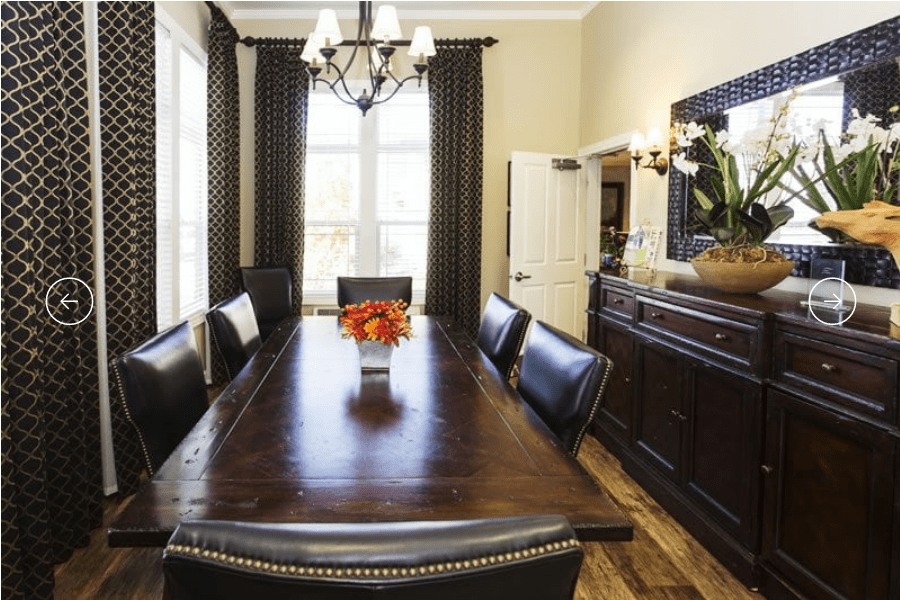 A beautifully set dining room with a large wooden table and upholstered chairs, featuring a chandelier and elegant curtains.