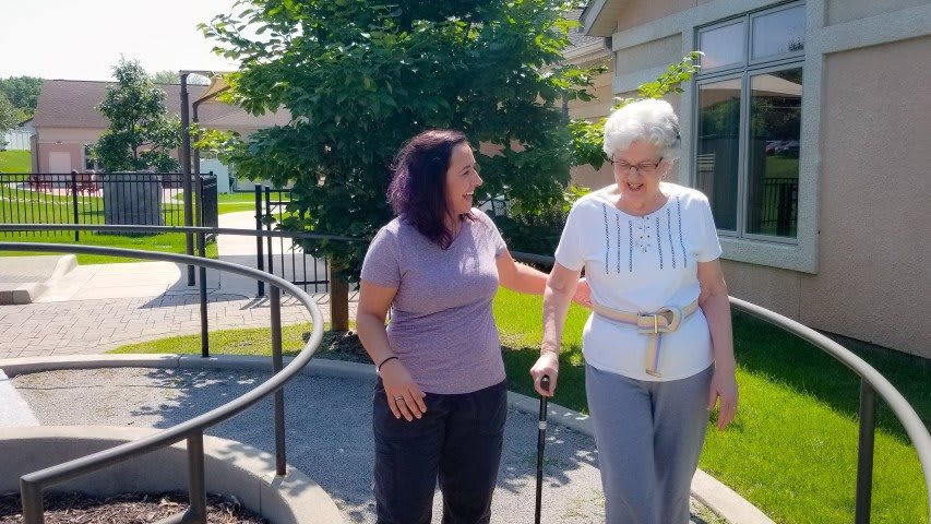 A caregiver assisting an elderly woman walking outdoors on a sunny day.