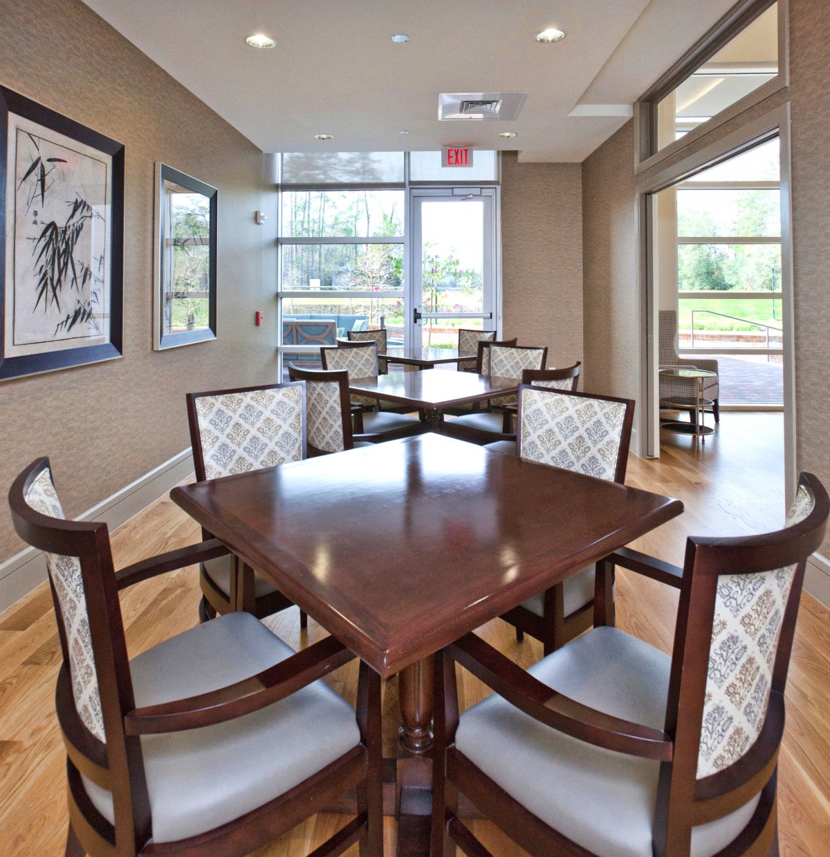 A modern dining area with a wooden table and cushioned chairs, featuring large windows and artwork on the walls.