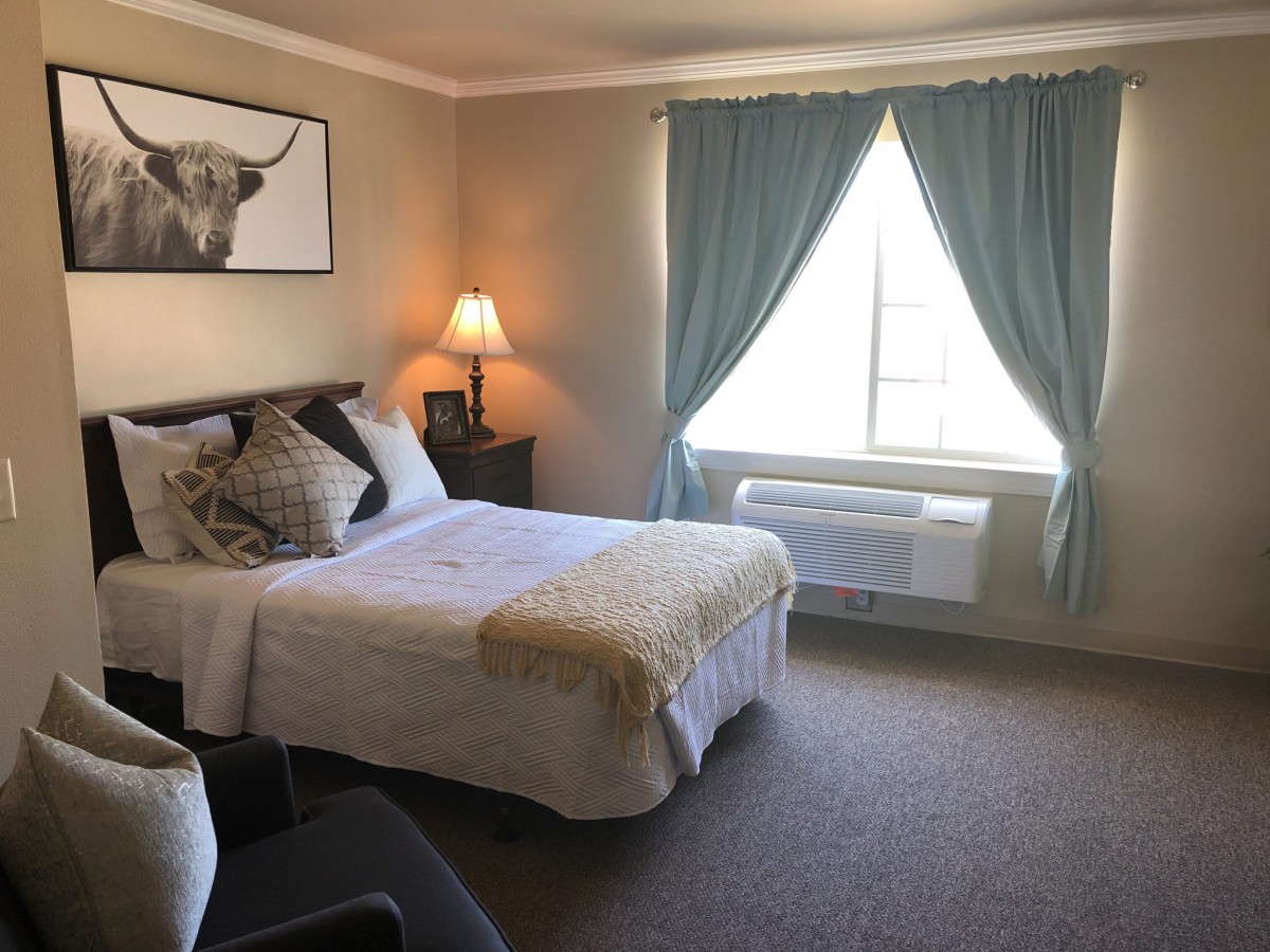 A cozy bedroom with a neatly made bed, soft lighting from a lamp, and large windows with blue curtains.