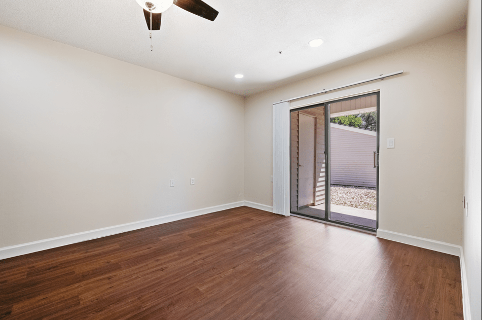 A spacious empty room with hardwood flooring, featuring a ceiling fan and a sliding glass door letting in natural light.
