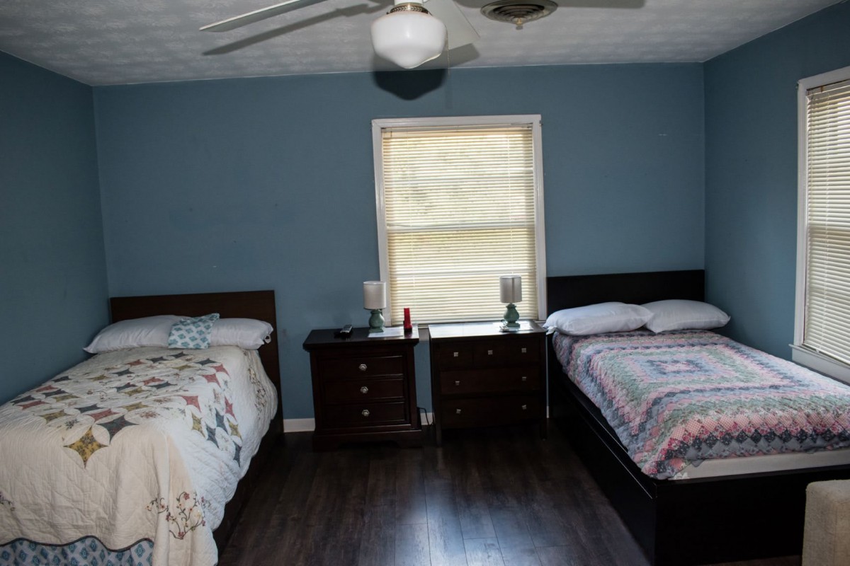 A bedroom featuring twin beds separated by a nightstand placed under a window with blinds, blue-painted walls, and a ceiling fan overhead.