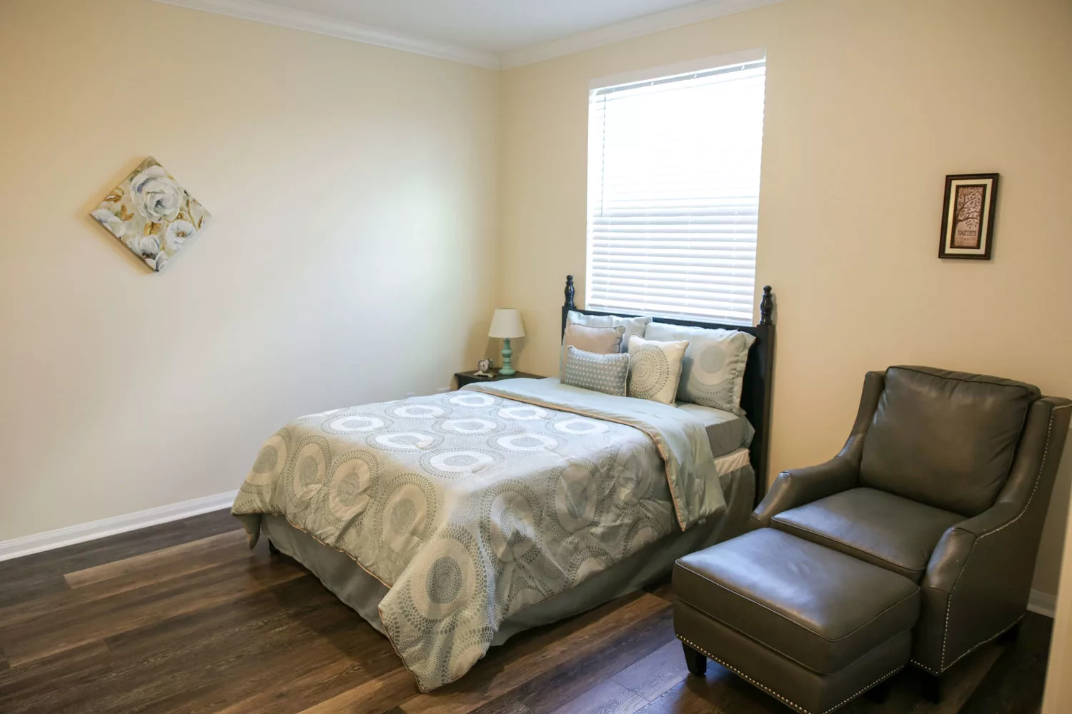 A cozy bedroom with a neatly made bed, upholstered chair, and soft lighting from a table lamp.