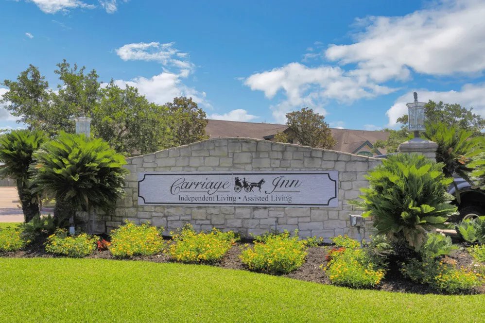 A welcoming neighborhood entrance sign set against a stone wall with well-maintained landscaping and a clear blue sky.