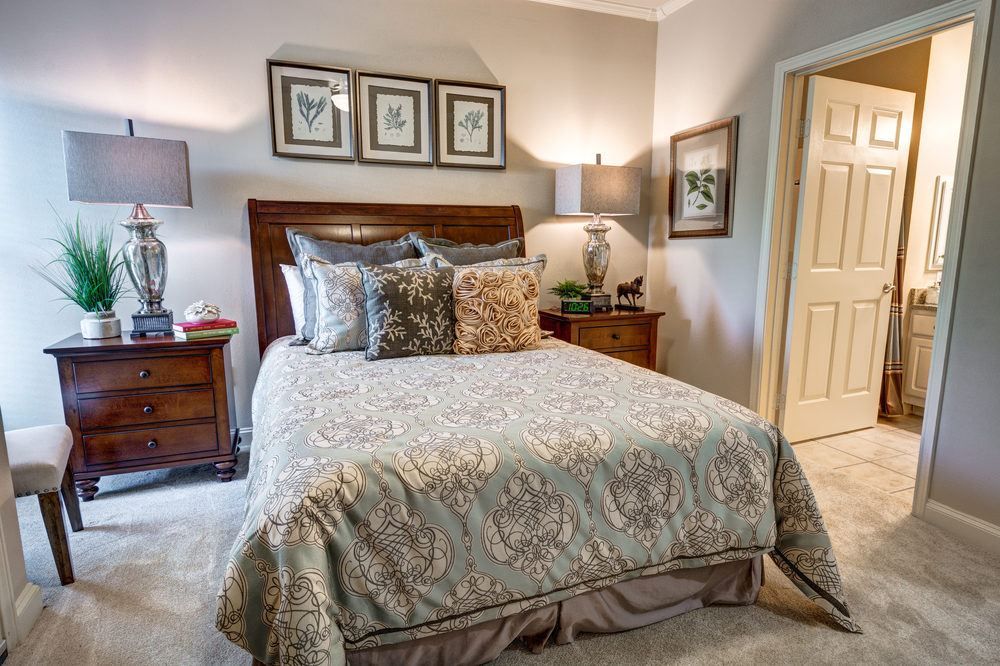 A cozy bedroom featuring a neatly made bed with multiple pillows, flanked by nightstands and lamps. The walls are adorned with framed artwork, creating a warm and inviting atmosphere.