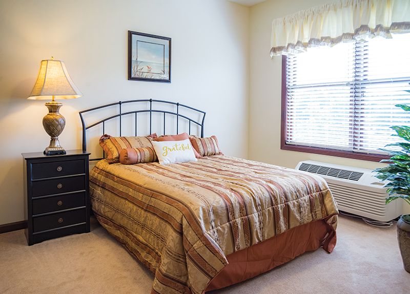 A cozy bedroom with a neatly made bed, a bedside lamp, and a window letting in natural light.