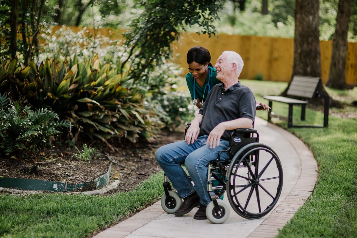 A caregiver assists an elderly person in a wheelchair along a garden path.
