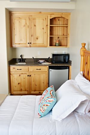 A small kitchenette setup next to a bed with wooden cabinets, microwave, and refrigerator.