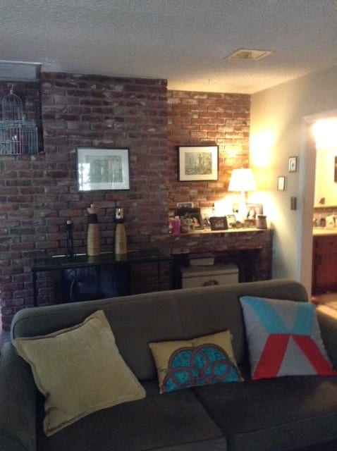 A cozy living room with an exposed brick wall, featuring a sofa adorned with decorative pillows and warm lighting from a table lamp.