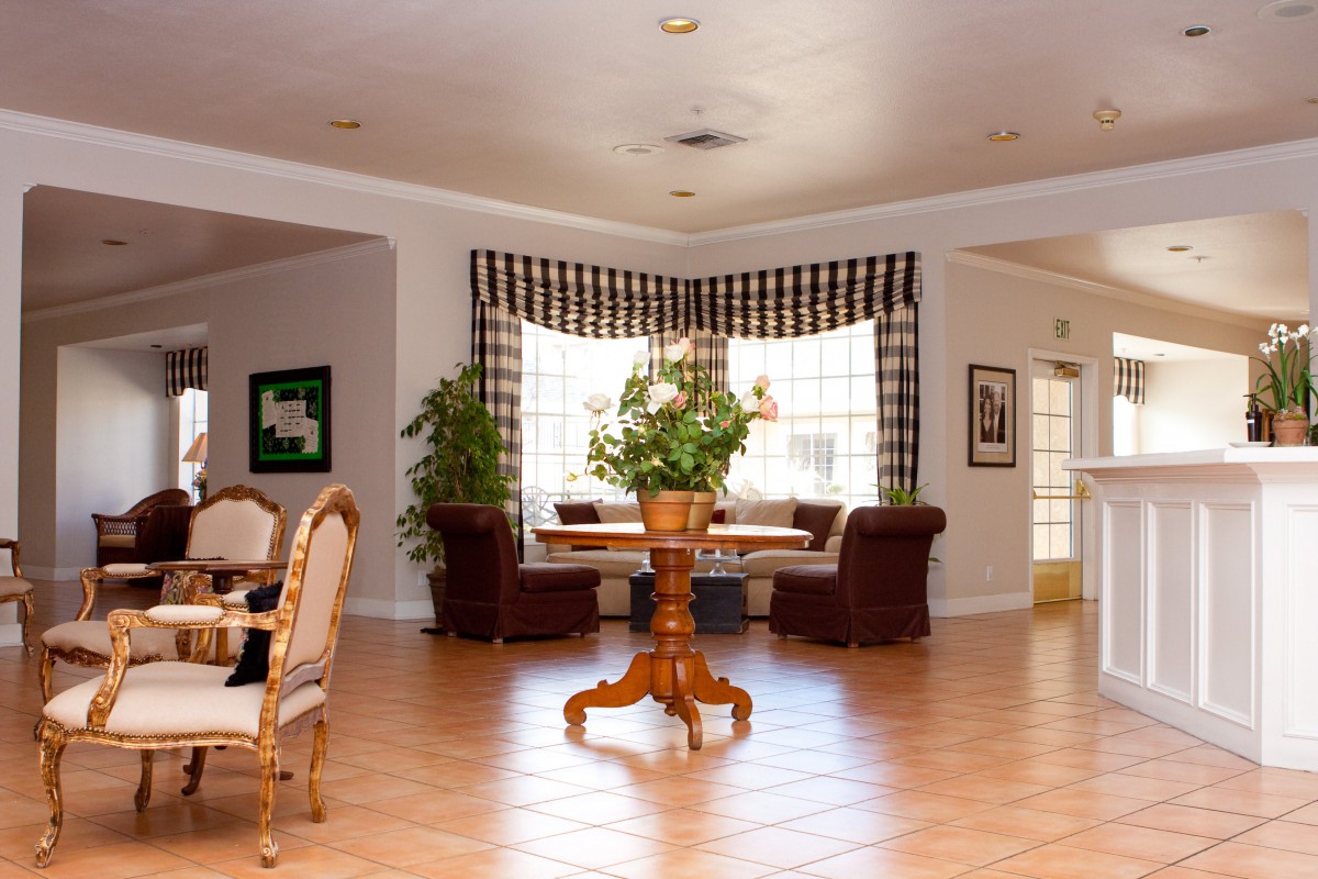 A well-lit living room with wooden floors, featuring a central table surrounded by chairs and green plants near the windows.
