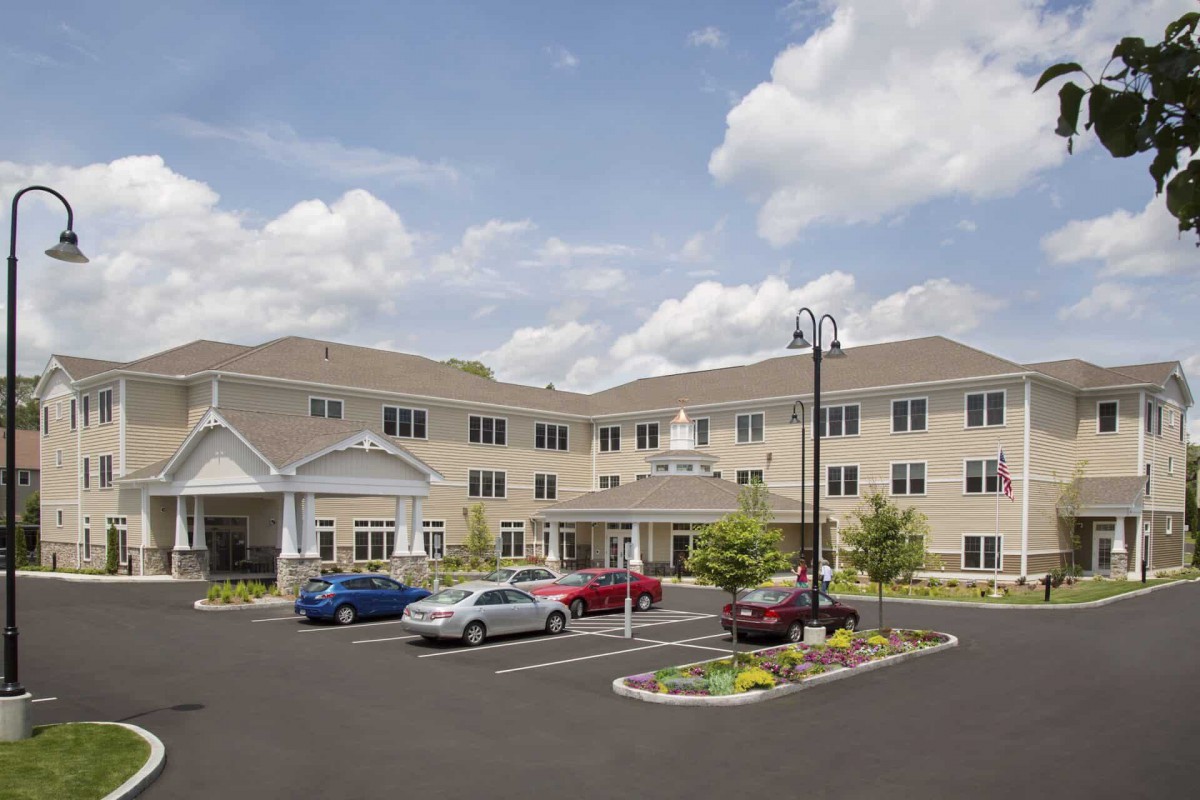 A multi-story building with a parking lot in front, featuring several parked cars and landscaped areas under a partly cloudy sky.