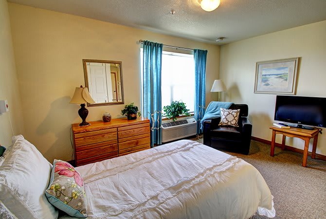 A cozy bedroom featuring a neatly made bed, a wooden dresser, an armchair by the window with blue curtains, and decorative plants.