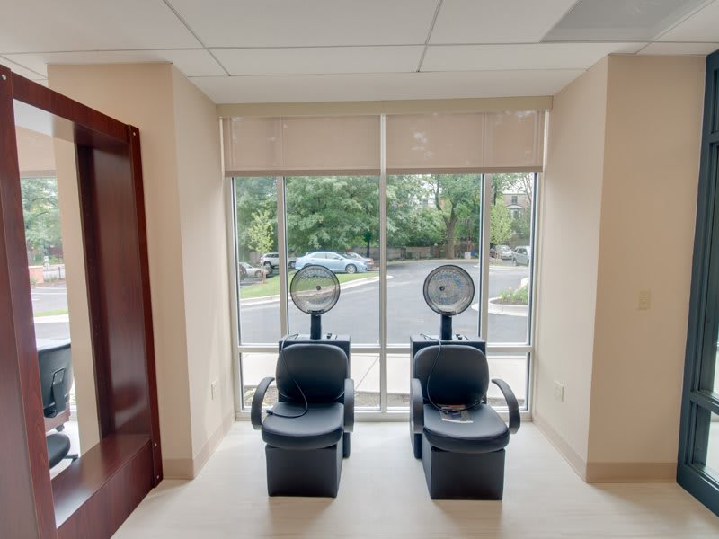 A bright salon interior with two chairs positioned under hair dryers near large windows.