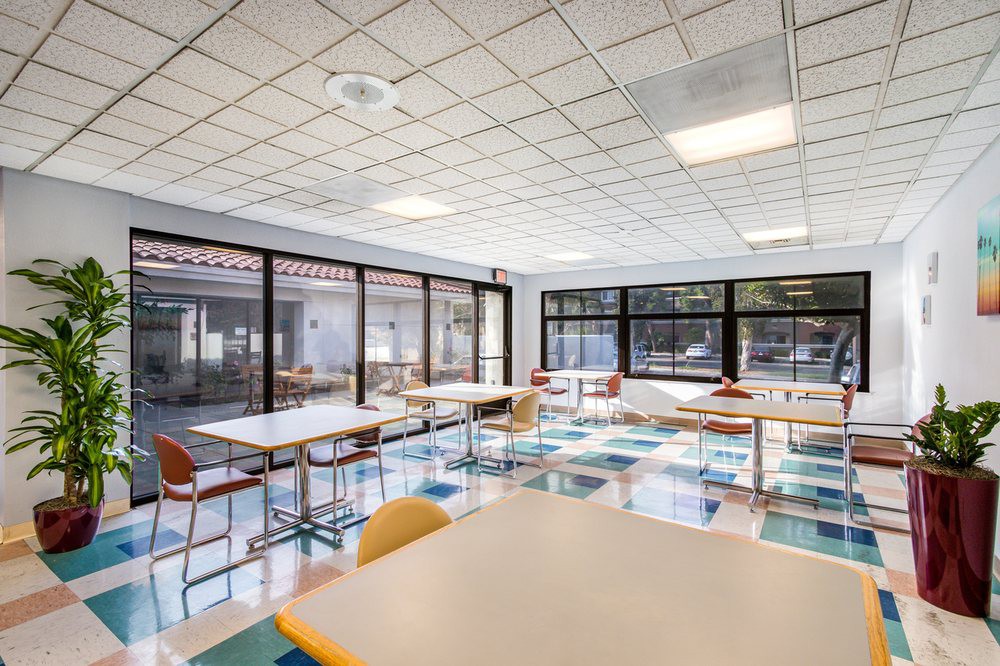 A bright classroom with tables and chairs arranged under large windows.