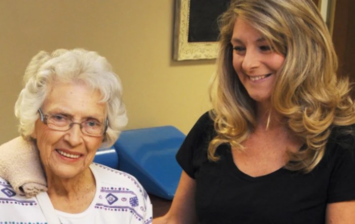 An elderly woman and a younger woman smiling together indoors.