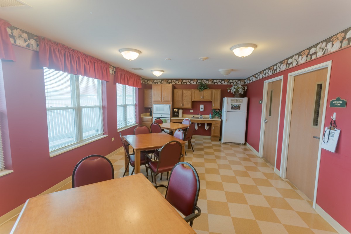 A kitchen and dining area with red walls, featuring multiple tables and chairs, a checkerboard floor, and several appliances.