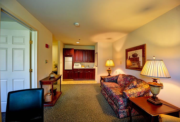 A cozy living room with a sofa, table lamps, and a view into a kitchenette with wooden cabinets.