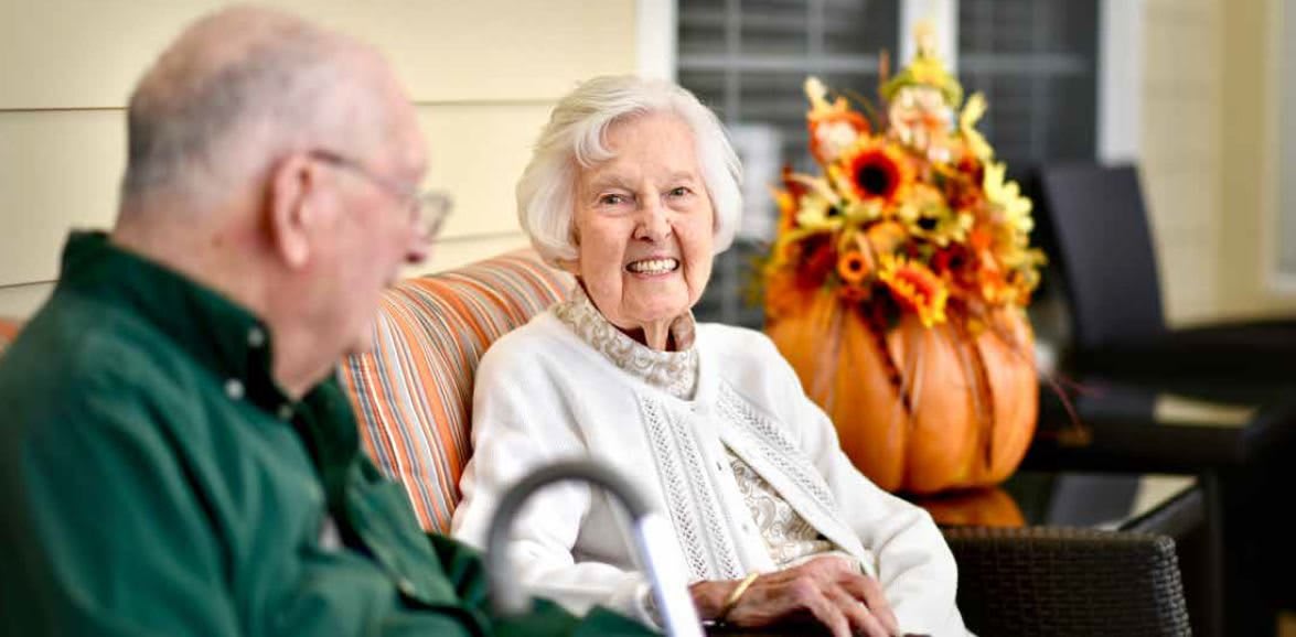 An elderly couple sitting outdoors, smiling and enjoying each other's company with autumn flowers in the background.
