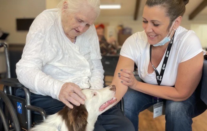An elderly woman and a caregiver enjoy spending time with a friendly dog indoors.
