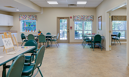 A bright and clean dining area with several tables and chairs, decorated with patterned curtains and ample natural light from large windows.
