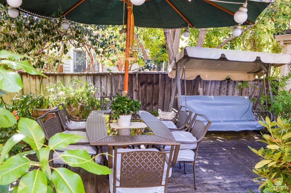 A cozy outdoor patio area with a table, chairs, and an umbrella surrounded by greenery.