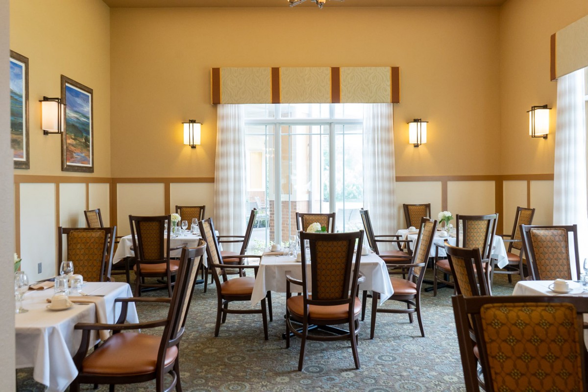 A well-lit dining area with arranged tables and chairs, featuring elegant lighting and large windows with curtains.