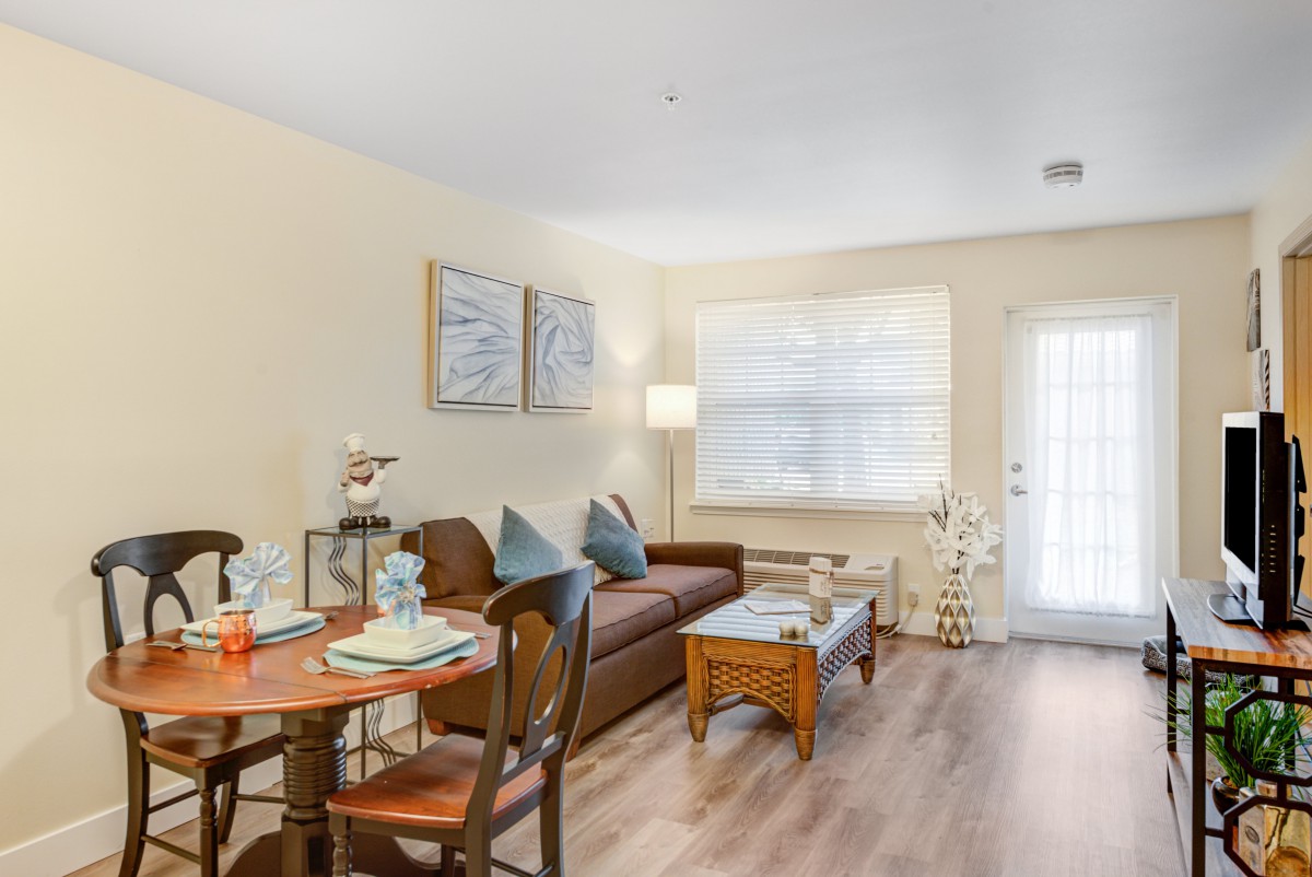 A cozy living room with a sofa, dining table, chairs, and a standing lamp near a window.