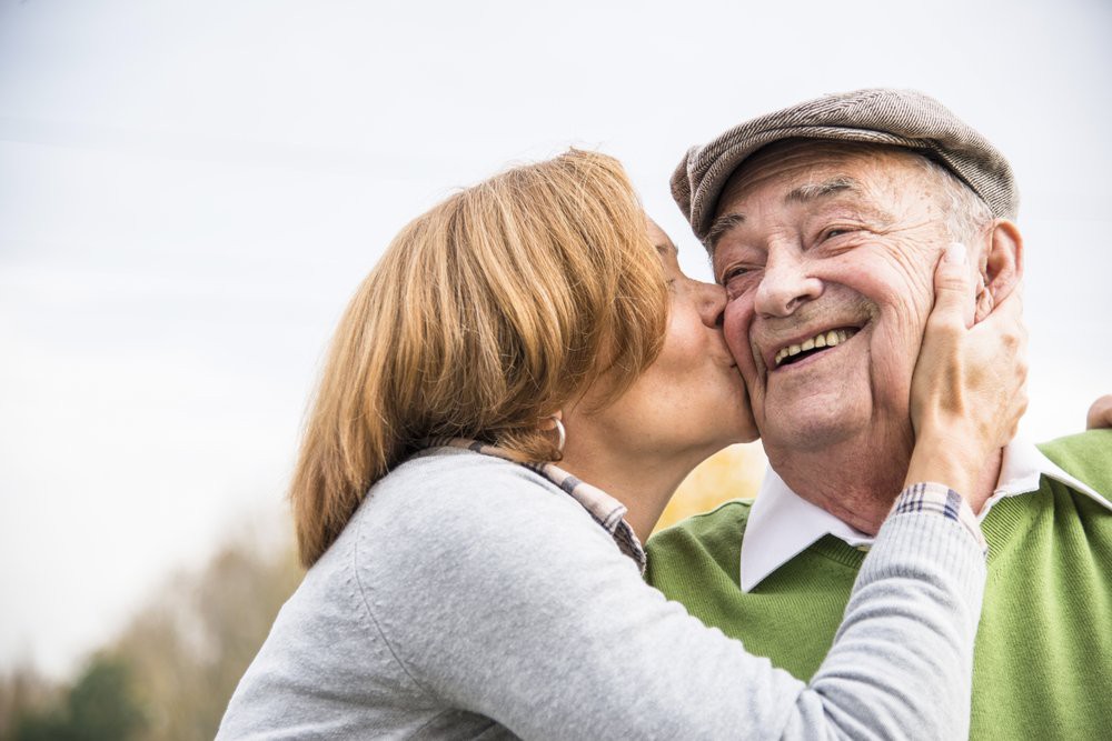 An elderly woman affectionately kisses an elderly man on the cheek, both smiling and enjoying a moment together outdoors.