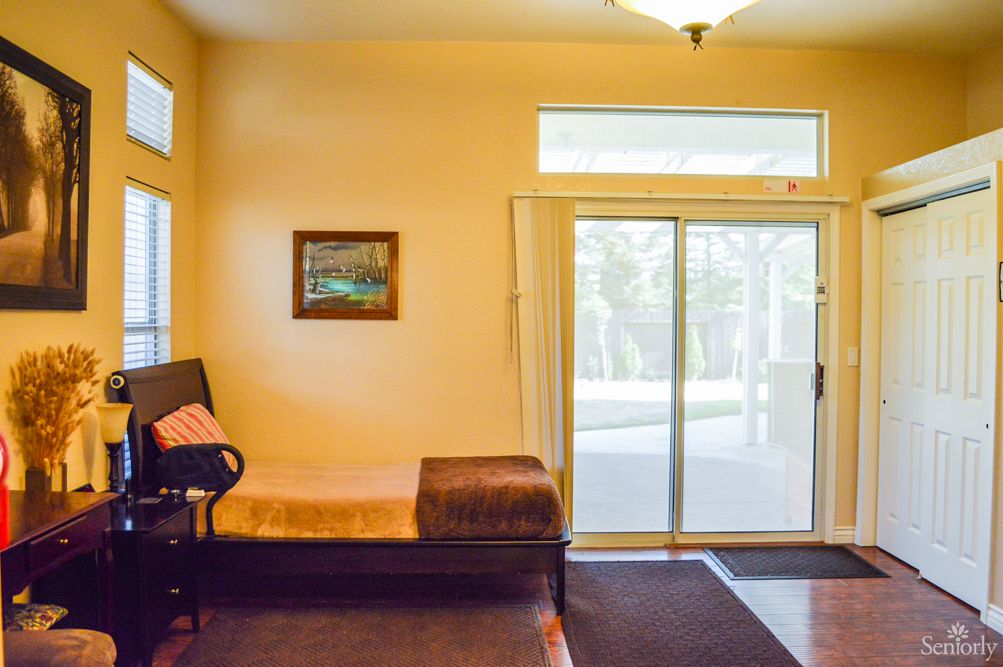 A cozy living room interior featuring a daybed next to a sliding glass door and window, with a painting on the wall and a carpet on the floor.