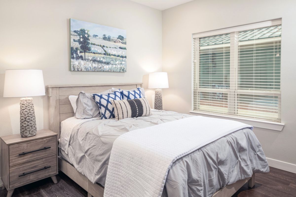 A cozy and well-decorated bedroom featuring a bed with pillows, a bedside lamp, and a scenic painting above the headboard.