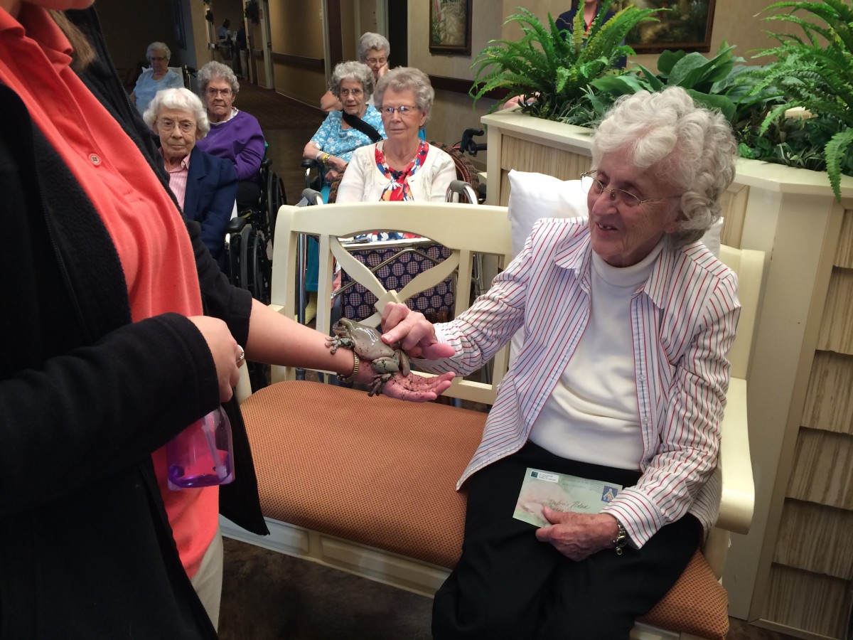 A seated elderly woman gently holds a small animal while interacting with another person in a senior living community.