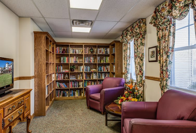 A cozy library corner with bookshelves, armchairs, and a small table with flowers.