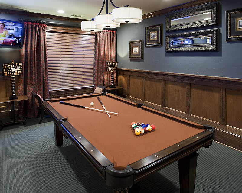 A stylish game room featuring a pool table with cue sticks and balls, surrounded by elegant wood paneling and lit by a chandelier.