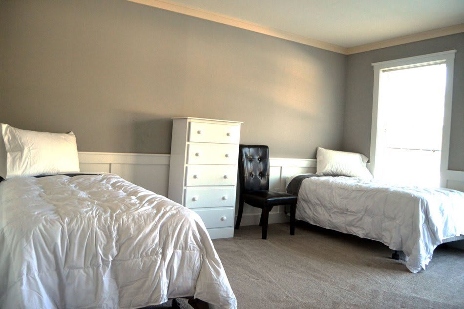 A minimalist bedroom with two beds, a dresser, and a chair in neutral tones.