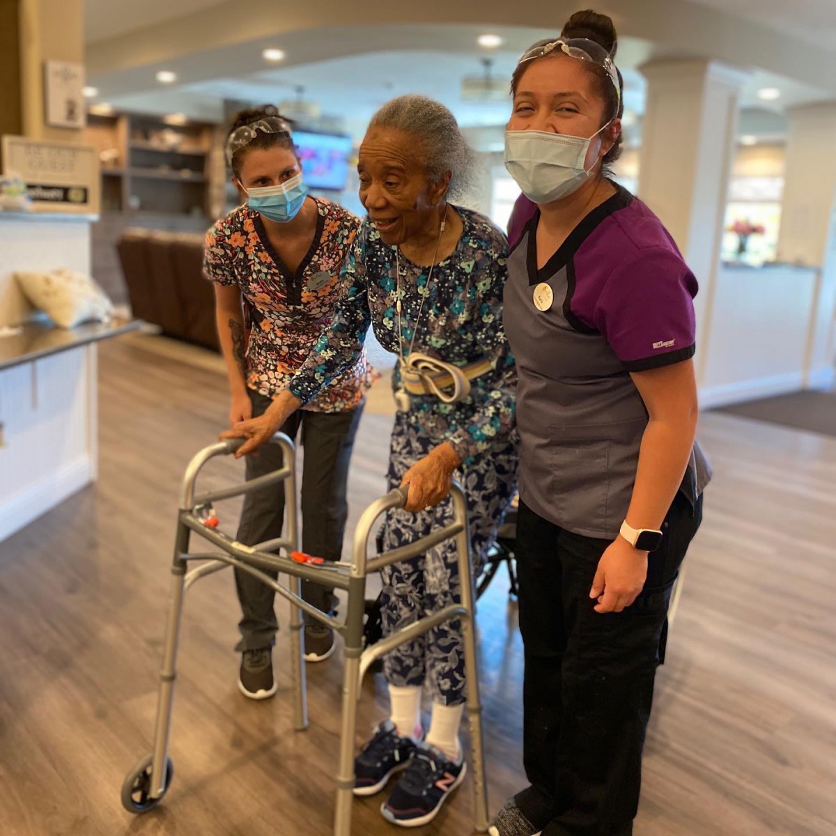 An elderly woman receives assistance from two healthcare workers while using a walker in a rehabilitation center.