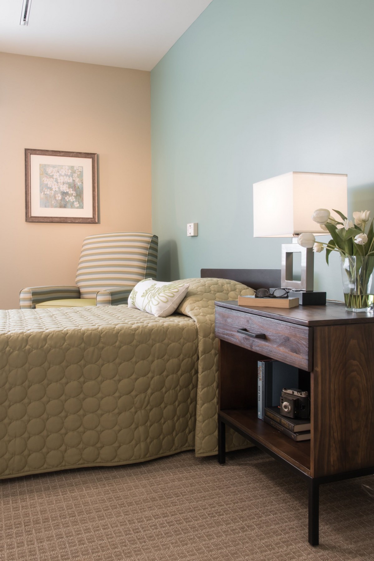 A cozy bedroom with a neatly made bed, a nightstand with a lamp and flowers, and a striped armchair.